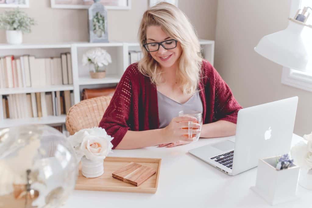 woman smiling holding glass mug sitting beside table with MacBook - fast ways of making money online