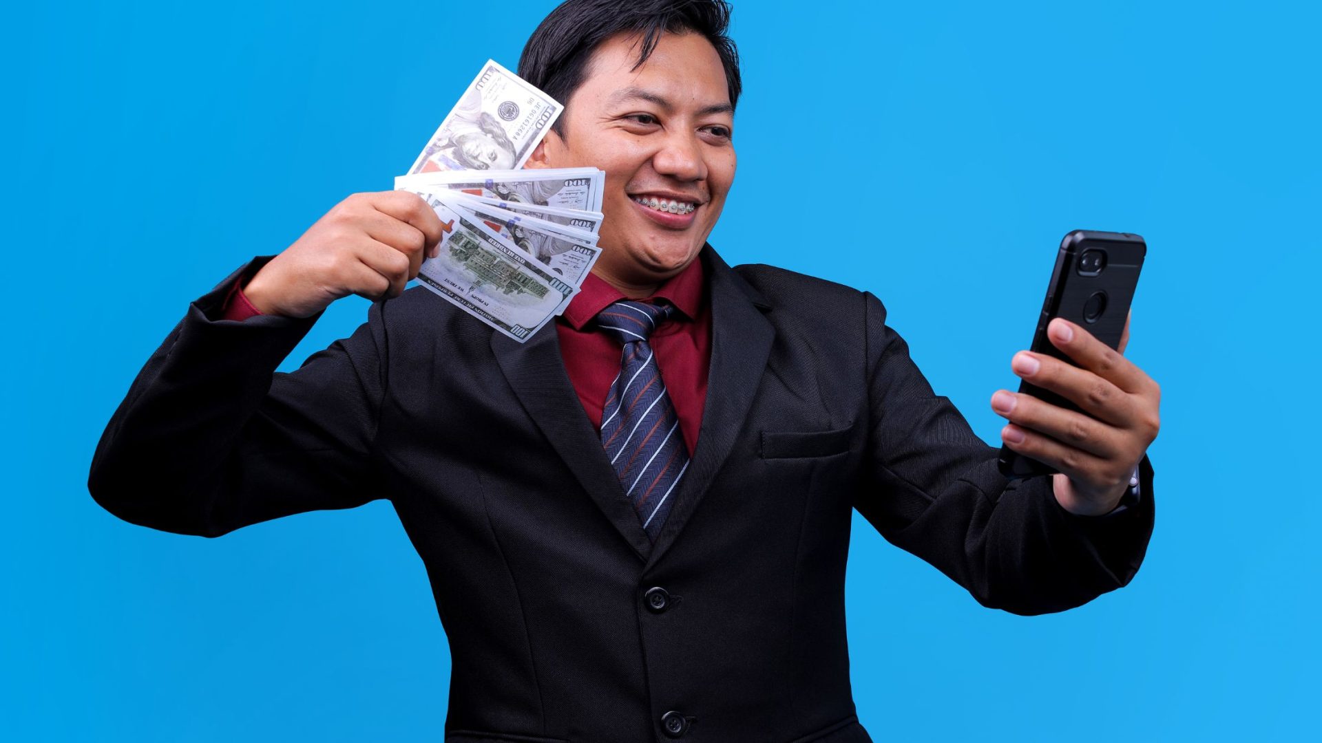 Get clients to pay faster. Asian man with money in one hand and a mobile phone in the other on blue background