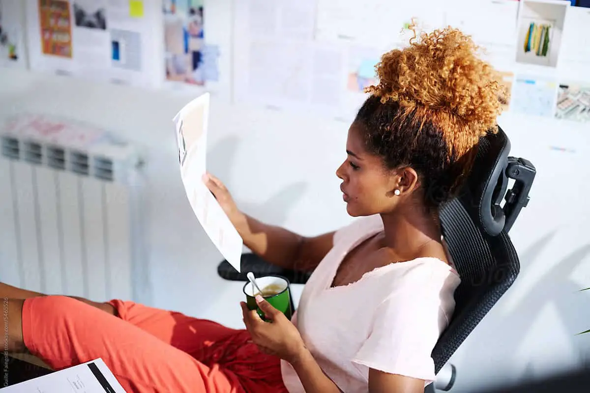 add resume to linkedin - Side view from above of focused entrepreneur woman with afro hair up read report CV, sit in comfortable office chair with mug of coffee in morning light. Relaxed woman sitting at offce