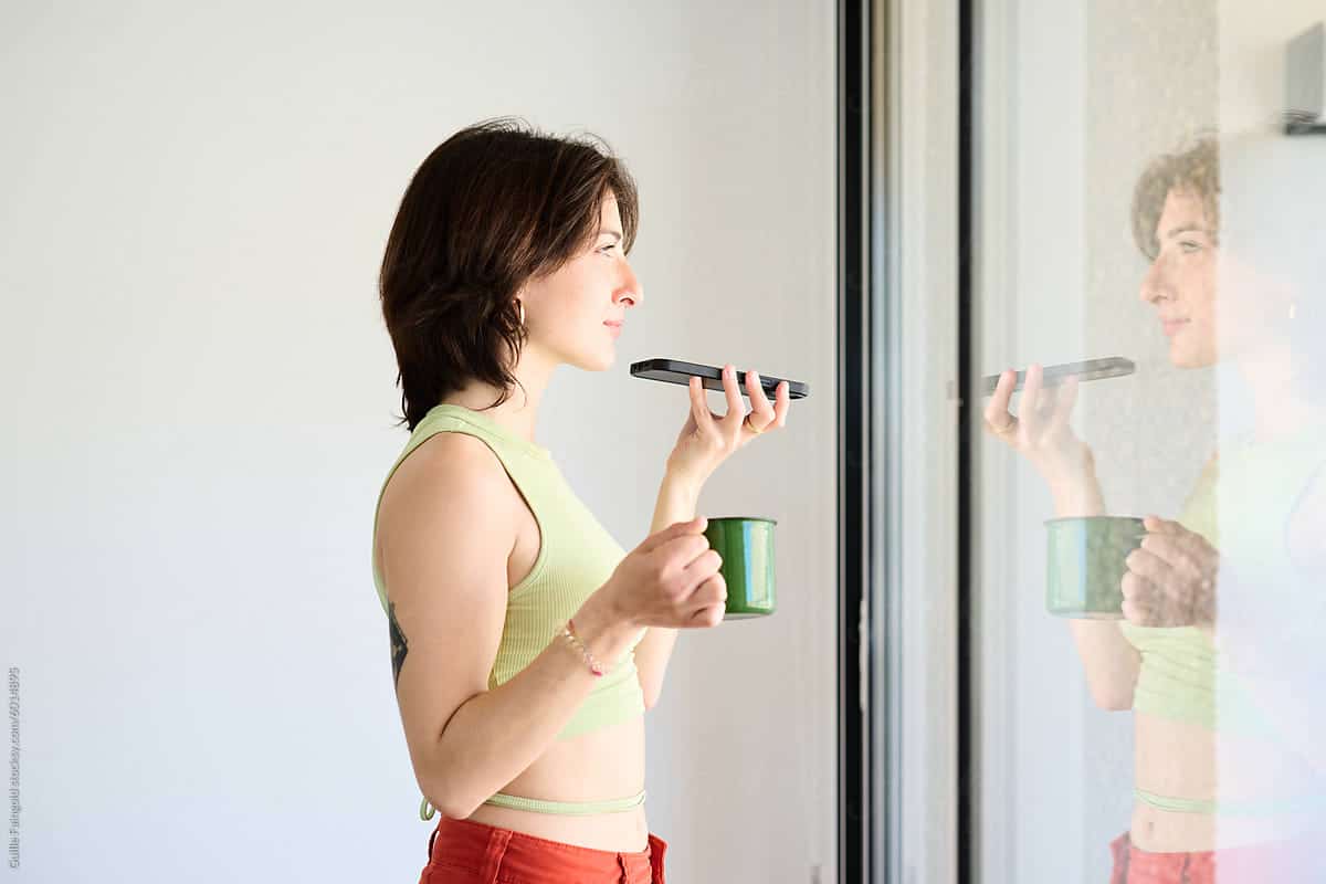 A young woman is seen happily recording a voice message on her smartphone, standing by a window holding a green cup at home. She is dressed casually, with her attention focused on the device, capturing a moment of modern communication. Zoho marketing automation