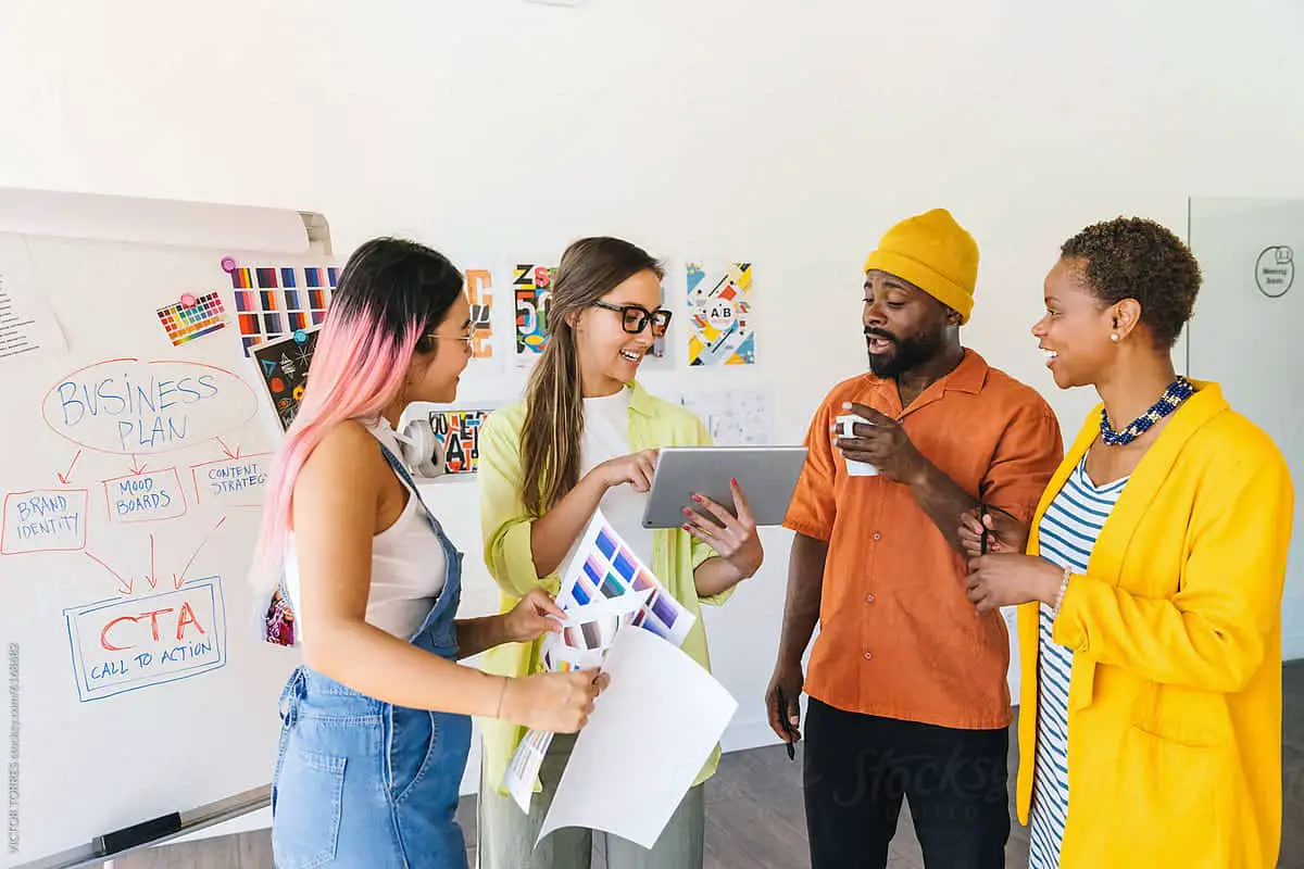 A diverse group of coworkers engage in a creative discussion around a business plan. They review color samples and notes, with a whiteboard displaying the plan and action steps in the background. Branding exercises