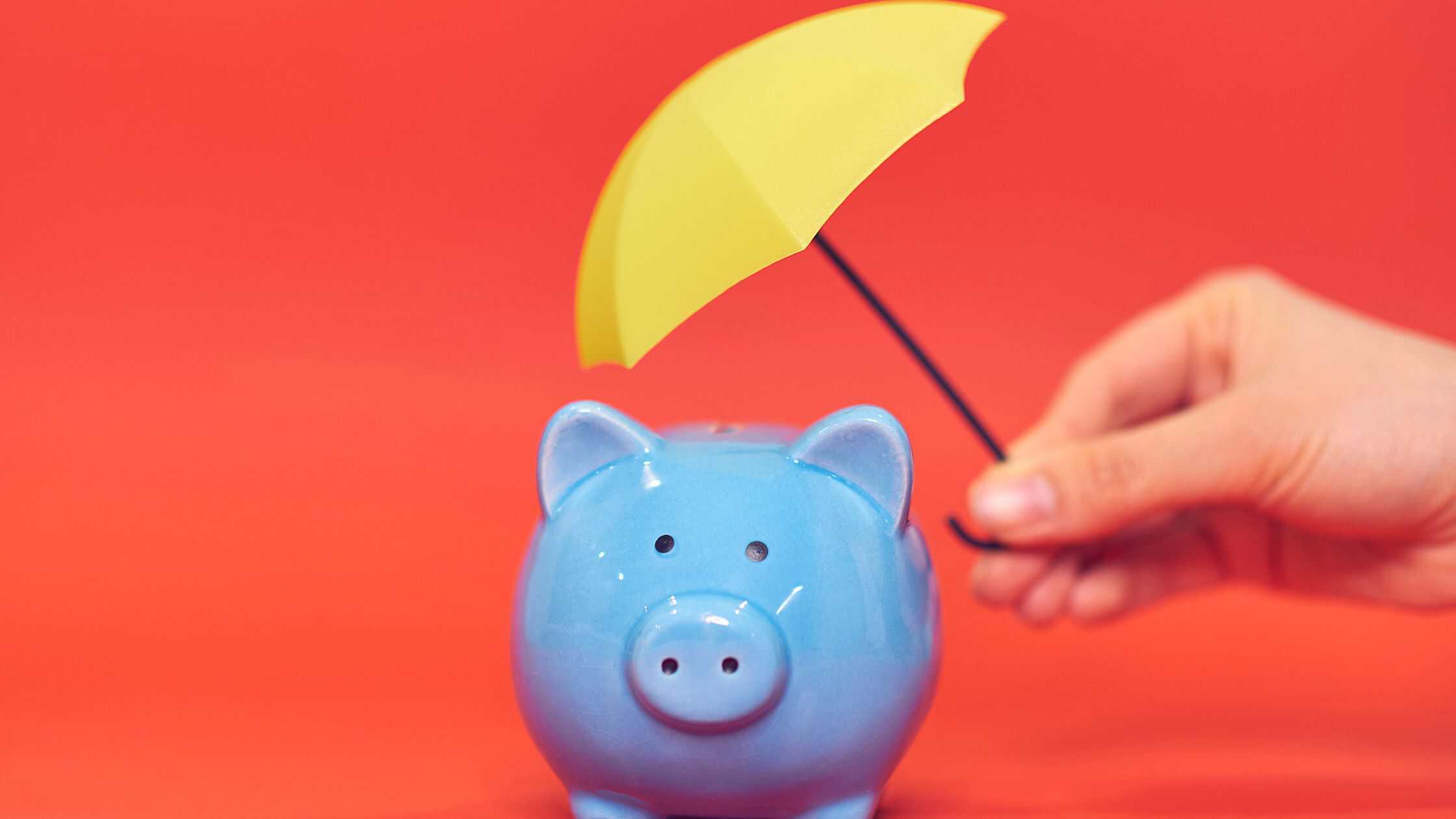 recession-proof small business. hand holding a small yellow umbrella protecting a blue piggy bank on red background